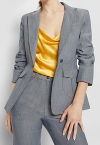 Blazer in denim con sottogiacca in satin giallo. Il blazer presenta un tessuto grigio, cuciture chiare e tasche inclinate. Design aderente.