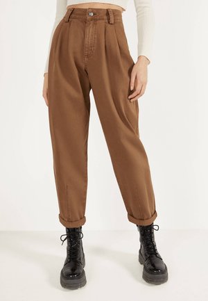 Brązowe spodnie z wysokim stanem o zwężanych nogawkach z podwiniętymi mankietami, noszone z czarnymi sznurowanymi butami typu combat i kremowym, prążkowanym topem z długim rękawem.