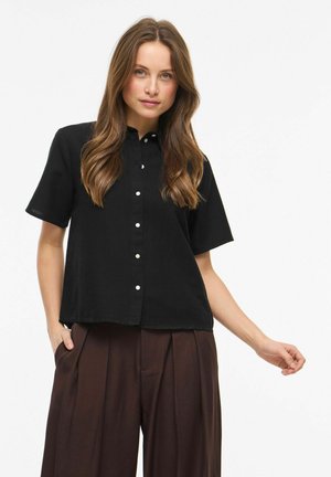 Jeune femme aux longs cheveux porte une chemise noire à manches courtes boutonnée et un pantalon marron ample, posant devant un fond blanc uni.