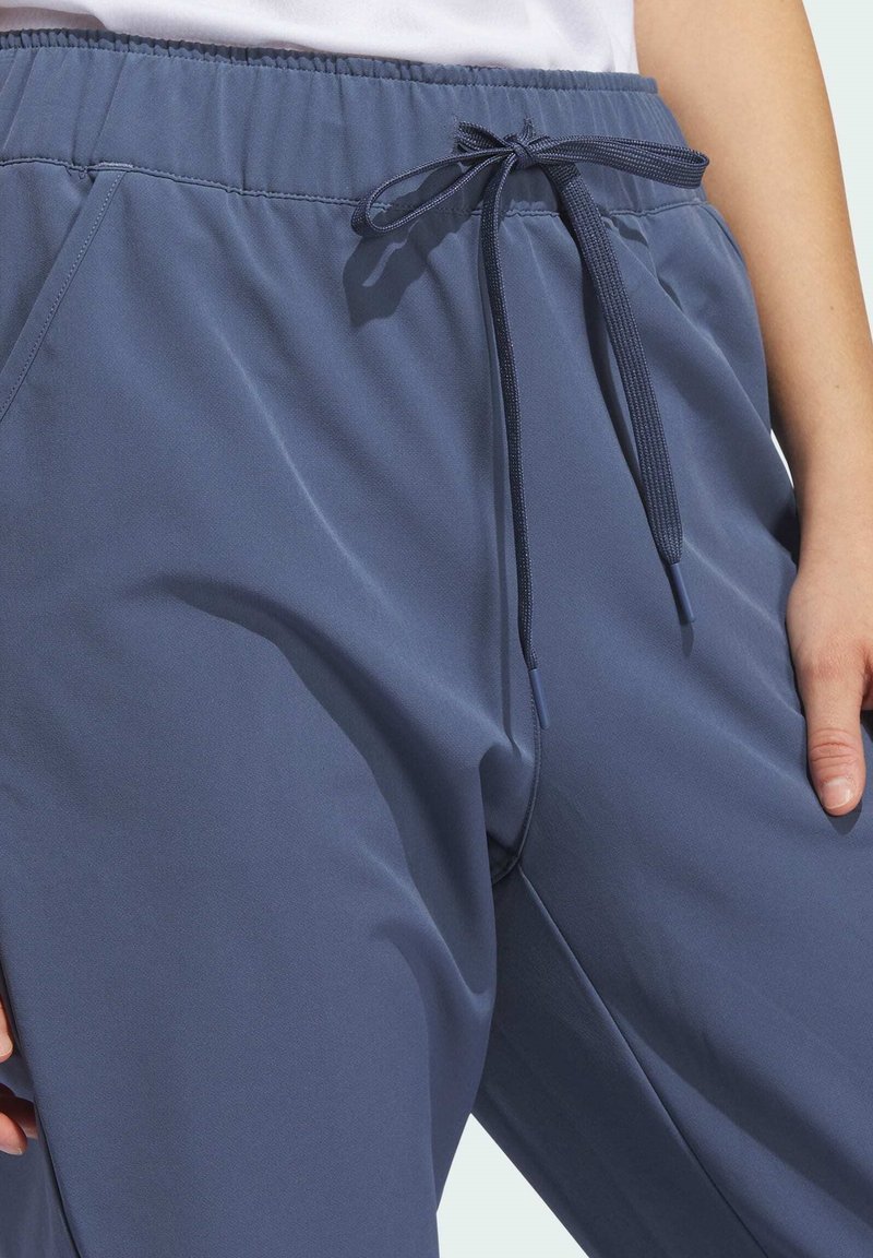 Gros plan sur une personne portant un pantalon élastique bleu à taille haute avec un cordon noué et des poches latérales, la main reposant près de la hanche.