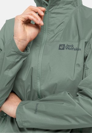 Veste imperméable vert clair en matière lisse, avec un col montant, une fermeture éclair intégrale, et les logos "Jack Wolfskin" et "Texapore" à l'avant.