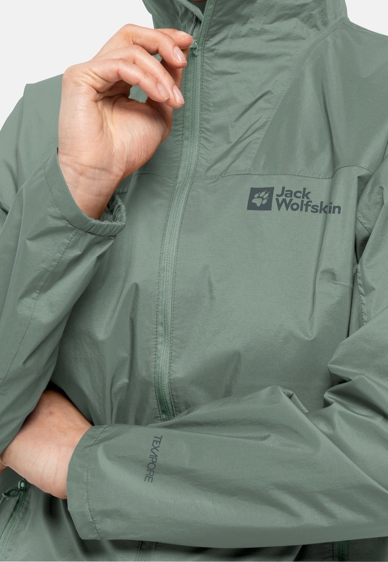 Chaqueta impermeable verde claro hecha de un material suave, con un cuello alto, cremallera completa y los logotipos "Jack Wolfskin" y "Texapore" en la parte delantera.