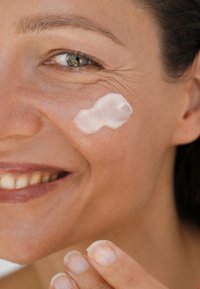 Zbliżenie na kobietę, która nakłada gładką, białą krem na policzek, prezentującą klarowną skórę o minimalnej teksturze i naturalnym, zdrowym blasku.