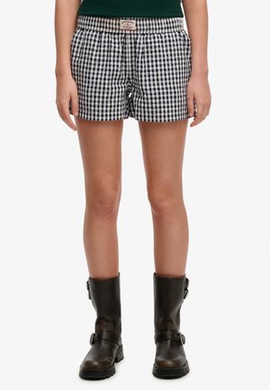 Person iført sort-hvide ternede shorts med Superdry-mærke, mørkegrøn top og mørkebrune spænde støvler stående oprejst.