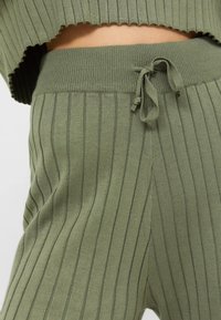 Groene geribbelde gebreide top met een golvende zoom, gecombineerd met bijpassende broek met koord, die verticale strepen en een zachte textuur toont.