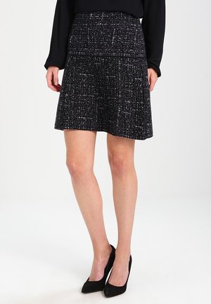 Jupe en tweed noir avec taille cintrée et ourlet évasé. Le tissu présente une texture subtile avec des éclats blancs. Portée avec des talons noirs.