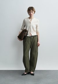 Jeune personne debout contre un mur gris, portant un cardigan crème boutonné, un pantalon ample olive, des ballerines noires pointues, tenant un sac en daim marron.