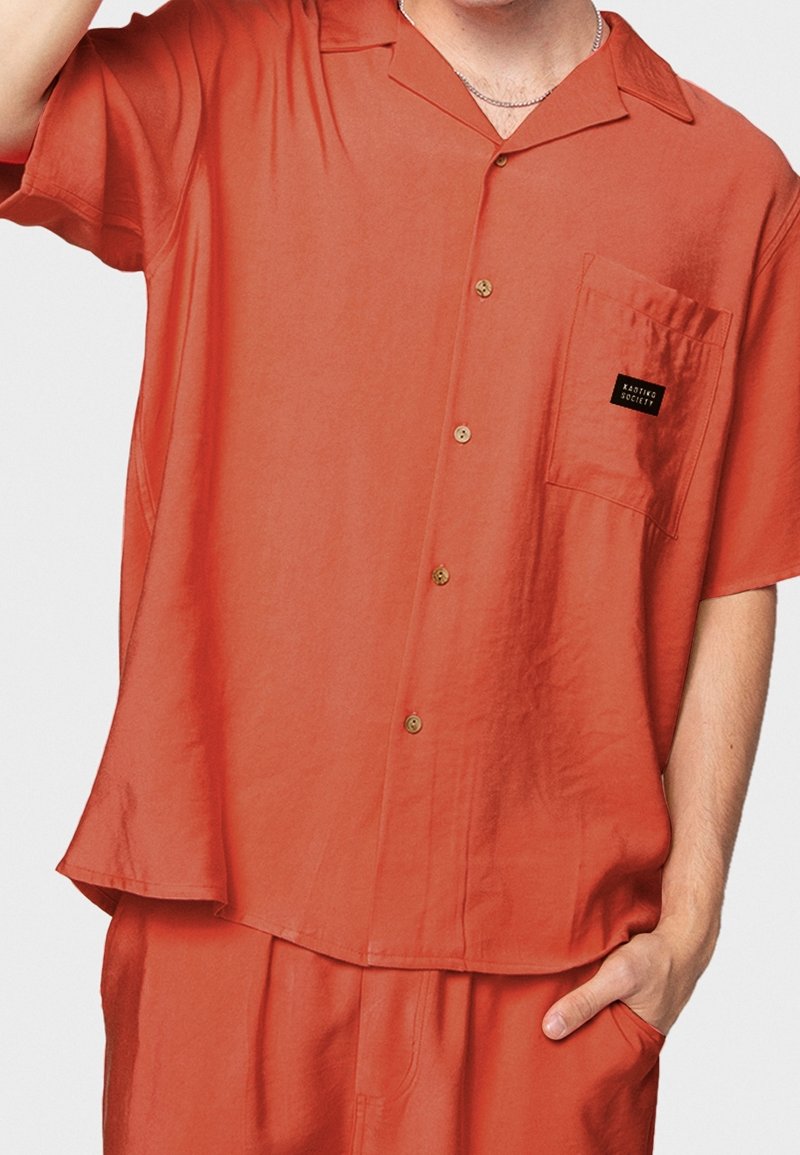 Korallfärgad kortärmad skjorta med knäppning och bröstficka, med en varumärkesetikett; tillverkad av mjukt tyg och har en avslappnad passform.