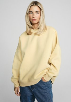Blondynka o prostych włosach, ubrana w luźną bladoróżową bluzę i niebieskie dżinsy, stoi z jedną ręką w kieszeni.