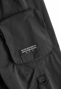 Schwarze Stoffjacke mit einer Fronttasche mit einem Etikett, strukturierten Oberfläche und Druckknopfverschluss. Auffällige Koordinaten auf dem Etikett aufgedruckt.