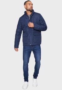 Marineblå quiltet jakke med høj krave, frontknaplukning og to sidelommer, parret med blå jeans og hvide sneakers.