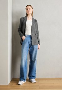 Blazer grigio a fantasia su una camicia bianca, abbinato a jeans a gamba larga blu chiaro e sneaker bianche, su uno sfondo neutro.