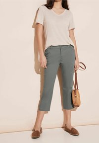 Kvinna iklädd en ljusbeige V-ringad kortärmad topp, grå capribyxor med ficka med dragkedja, bruna slip-on skor och håller en rund stråväska.