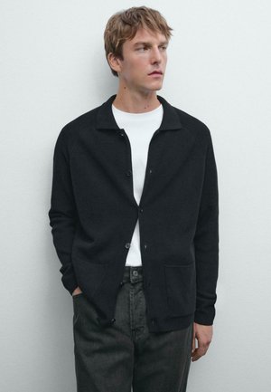 Cardigan a maglia scura con colletto, bottoni frontali e due tasche, indossato sopra una camicia bianca. Il modello posa su uno sfondo neutro.