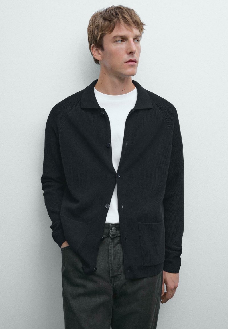 Donkere gebreide cardigan met een kraag, voorknopen en twee zakken, gedragen over een wit shirt. Model poseert tegen een neutrale achtergrond.