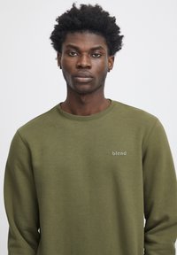 Jonge man met krullend haar, gekleed in een groene sweatshirt met ronde hals met het logo "blend" op de borst, tegen een effen witte achtergrond.