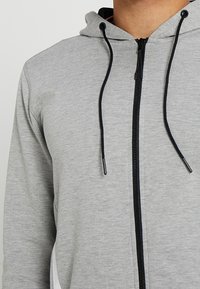 Sudadera con cremallera gris con cremallera y cordones negros. Presenta una textura de tela suave y acentos deportivos en los laterales. Capucha adjunta.