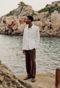 Un homme se tient sur un rivage rocheux, portant une veste blanche, un pantalon marron et des sandales, regardant sur le côté avec de l'eau et des falaises escarpées en arrière-plan.