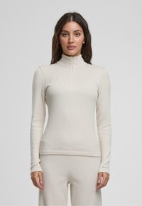 Lys beige zip-neck langermet topp i ribbestrikket stoff, tettsittende design, med en jevn tekstur og subtile detaljer ved mansjettene.