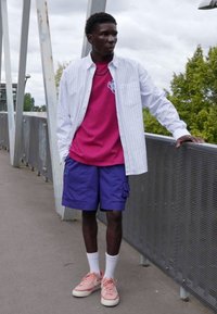 Hvid stribet skjorte med knap, lyserød t-shirt med grafik, lilla cargo shorts, lyserøde sneakers, står på en bro med grønne omgivelser i baggrunden.