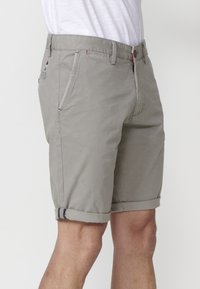 Shorts grii deschis dintr-un material texturat, având margini rulate, buzunare laterale și un detaliu mic în roșu și albastru.