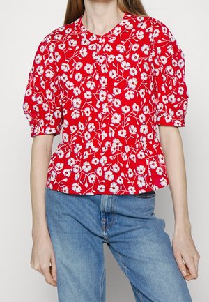 Žena v červené blůze s bílým květinovým vzorem, nafukovacími krátkými rukávy a knoflíky, doplněná světle modrými džínami.
