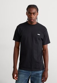 Zwarte katoenen t-shirt met een ronde hals en korte mouwen, voorzien van een klein wit "OBEY" logo op de linkerkant van de borst. Draag het met blauwe jeans.