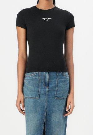 Person iført en tætsiddende sort T-shirt med korte ærmer med teksten "Tommy Jeans New York" og et blåt denimsnederdel med forlommer og en forflænse.
