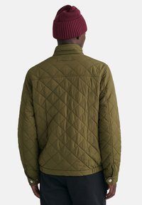 Giacca verde oliva trapuntata con colletto alto, polsini con bottoni a pressione e dettaglio dell'etichetta sul retro. Include un berretto di maglia maroon a coste.