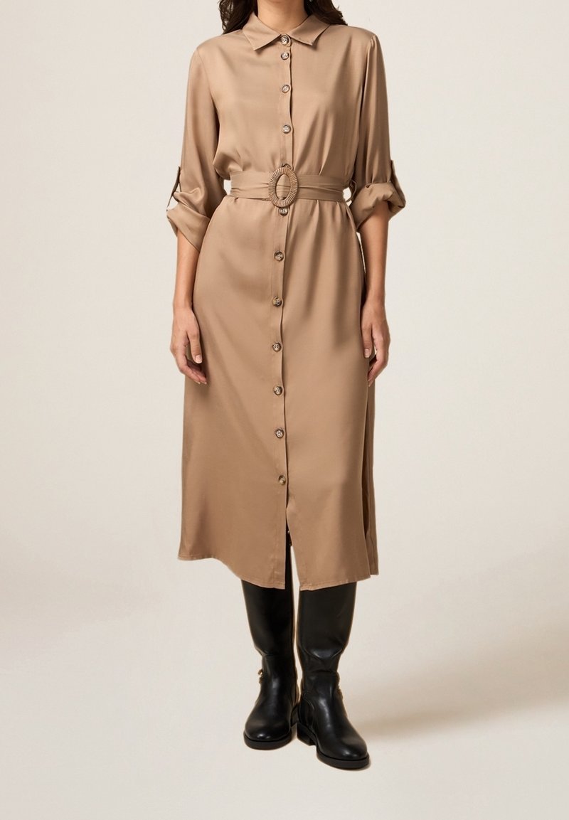 Femme portant une longue robe beige boutonnée, manches retroussées et ceinture assortie, associée à des bottes en cuir noires montant jusqu'aux genoux.