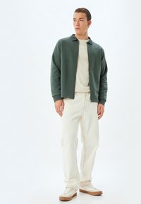 Chaqueta de suéter en poliéster verde oscuro, diseño con cuello, combinada con jeans de algodón crema y zapatillas beige. Modelo de pie contra un fondo claro.