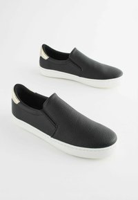 Fekete slip-on cipők texturált bőrből, fehér gumitalppal és fémes arany díszítéssel a saroknál. Minimalista dizájn.