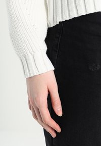 Bílý pletený svetr s žebrovanými manžetami, kombinovaný s černými denimovými kalhotami. Ruka viditelná na obrázku jemně spočívá podél bočního švu kalhot.