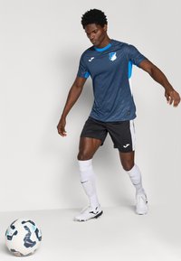 Maxi a maniche corte di colore blu scuro, con accenti di blu più chiaro, abbinato a pantaloni corti neri e calze bianche, accanto a un pallone da calcio con motivo.