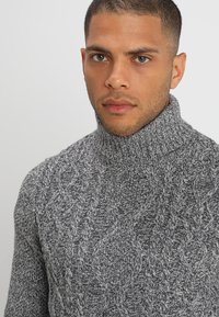 Suéter de cuello alto de punto gris con un patrón de cable texturizado y cuello acanalado. La tela tiene un aspecto suave y tejido con tonos grises variados.