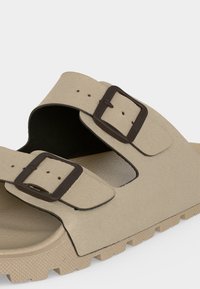 Sandale beige avec deux boucles rectangulaires réglables marron foncé et semelle texturée, présentée sur fond transparent.