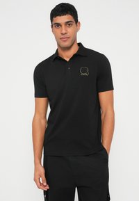 Čierne polokošeľa z bavlny, s krátkymi rukávmi a zlatou grafikou na ľavej strane hrudníka. Hladká textúra, klasický pohodlný strih.