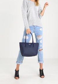 Bolsa de tela azul marino con dos asas, textura suave, que presenta un pequeño logo. El modelo lleva puestos unos jeans rasgados de color azul claro y un suéter gris.