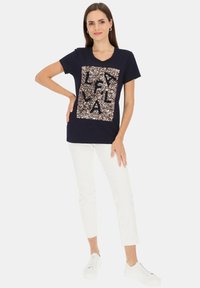 Granatowy bawełniany t-shirt z dekoltem w serek, ozdobiony złotym cekinowym kwadratem z czarnymi literami. W zestawie z białymi spodniami 7/8 i białymi trampkami.