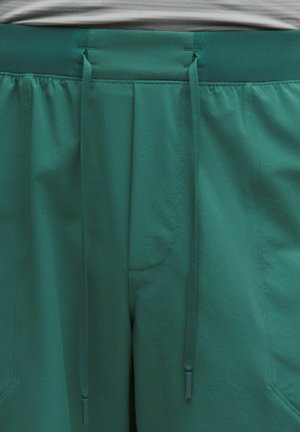 Zaļas teksturētas sporta bikses ar elastīgu vidukļa jostu, divām priekšējām kabatām un aukliņām, kas karājas centrā.