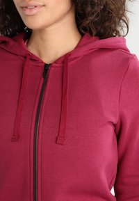 Bordová mikina se zipem a kapucí na stažení, vyrobená z texturované látky, s černým zipem a viditelným šitím podél švů.