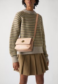 Crossbody torbica iz meke bež usnje z zaobljenim poklopcem in zlatimi dodatki. Ima en trak in čiste, preproste linije.