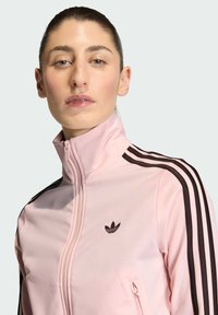 Ružová športová bunda s vysokým golierom, s dvojakým zipsom a hnedými tromi prúžkami. Obsahuje vyšívané logo na ľavej strane hrude.