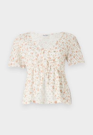 Blomstret bluse i let stof med hvit baggrund, der har rosa og grønne blomster, korte ærmer og en V-udskæring med en slipsedetalje.