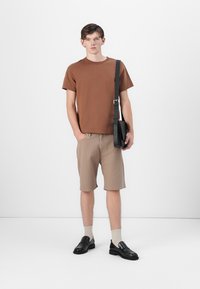 Bruine katoenen t-shirt, beige gestreepte shorts, zwarte leren loafers met sokken en een zwarte schoudertas. Model staat tegen een effen witte achtergrond.