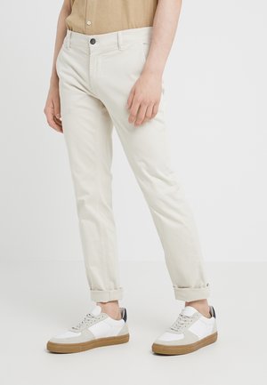 Mann trägt hellbeige, hochgekrempelte Chinos und weiße Sneaker mit Gummisohlen vor einem schlichten hellen Hintergrund.