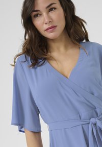 Vestido envolvente azul claro con mangas cortas y fluidas, escote en V y cinturilla atada. Textura suave con un ajuste relajado.