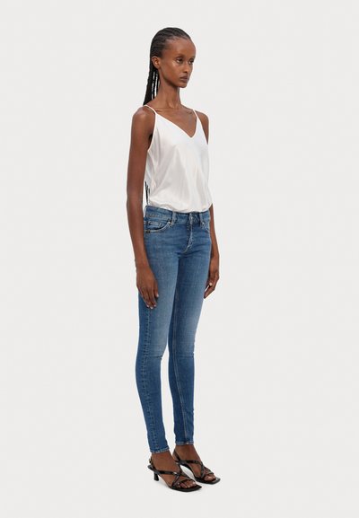 Jean skinny bleu en denim, associé à un cami en satin blanc. Porté avec des talons noirs à lanières, mettant en valeur une silhouette ajustée.