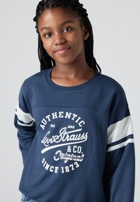 Jong meisje met vlechten, gekleed in een blauwe Levi Strauss sweatshirt met witte strepen op de mouwen en bedrukte tekst op de voorkant.