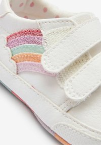 Baskets blanches avec des bandes Velcro, présentant un design multicolore pailleté en arc-en-ciel sur le côté. Semelle blanche texturée et doublure à pois.